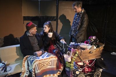 Kobieta i mężczyzna przykry kocem siedzą na ławce. Kobieta trzyma kubek , a drugą ręką wzóek dziecięcy wypełniony różnymi przedmiotami. Obok nich stoi mężczyzna.