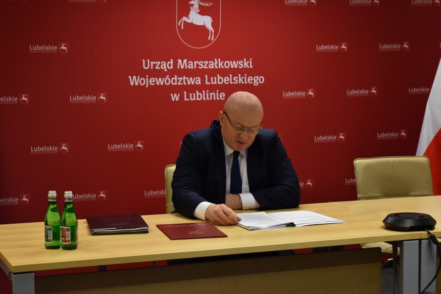 Przy stole nad dokumentami siedzi marszałek Stawiarski. W tle czerwona ścianka z herbem województwa i napisem Urząd Marszałkowski Województwa Lubelskiego w Lublinie.