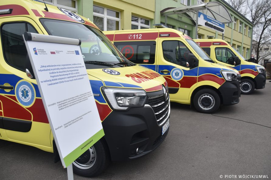 Trzy karetki pogotowia ratunkowego w żółtym malowaniu z niebiesko-czerwonymi pasami, ustawione jeden za drugim (widok na przód samochodów). W tle budynek Pogotowia Ratunkowego. Na pierwszym planie z lewej tablica z nazwą projektu unijnego.