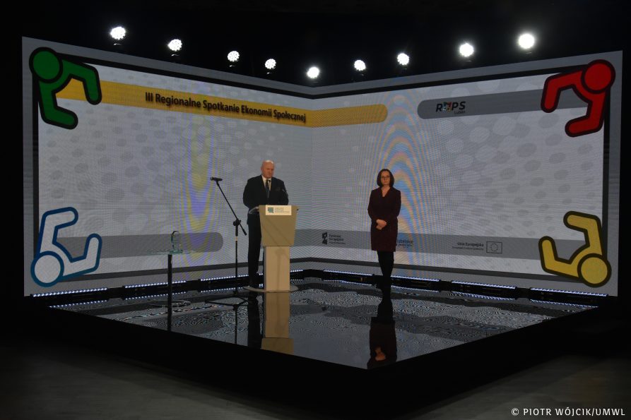 Szeroki kadr, na scenie ułożonym w romb przy mównicy stoi marszałek województwa Jarosław Stawiarski, z prawej dyrektor Regionalnego Ośrodka Pomocy Społecznej Małgorzata Romanko. W tle ekran wyświetlający ozdobne grafiki oraz nazwę wydarzenia