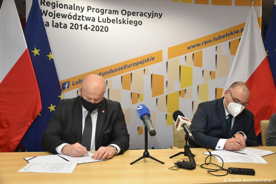 Marszałek Jarosław Stawiarski (z lewej) oraz dyrektor Michał Jedliński podpisują umowę na dofinansowanie. W tle ścianka Regionalnego Programu Operacyjnego, po obydwu stronach flagi Polski i Unii Europejskiej.
