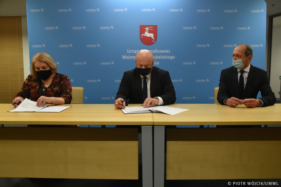 Przy stole siedzą od lewej Ewa Zybała Starosta Lubartowski, Jarosław Stawiarski Marszałek Województwa Lubelskiego, Lucjan Mileszczyk Wicestarosta Lubartowski. Starosta i Marszałek w trakcie podpisywania umowy. W tle niebieska ścianka z herbem województwa i logotypem lubelskie.