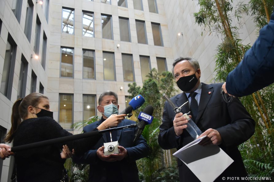 Konferencja prasowa, na zdjęciu po środku Konsul Generalny Ukrainy w Lublinie Vasyl Pavluk, wicemarszałek Zbigniew Wojciechowski. Panowie trzymają w rękach sprzęt medyczny. Wokół mikrofony i dziennikarze. W tle roślinność i fragment wnętrza urzędu marszałkowskiego. Widok lekko z dołu.