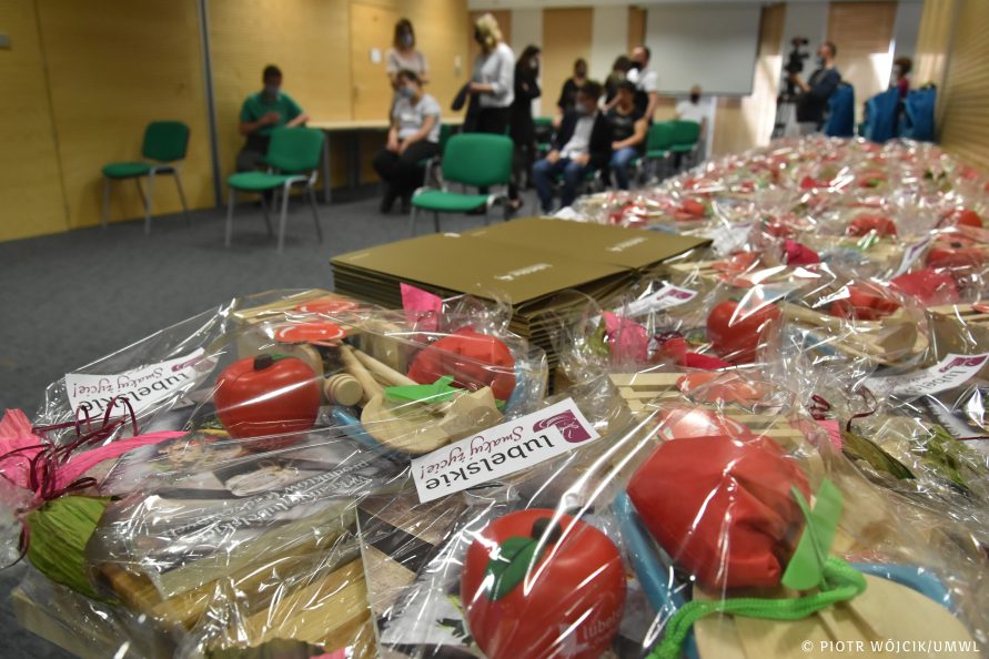 na stole leżą zestawy prezentowe zapakowane w przeźroczystą folię. W tle poza polem ostrości część laureatów.