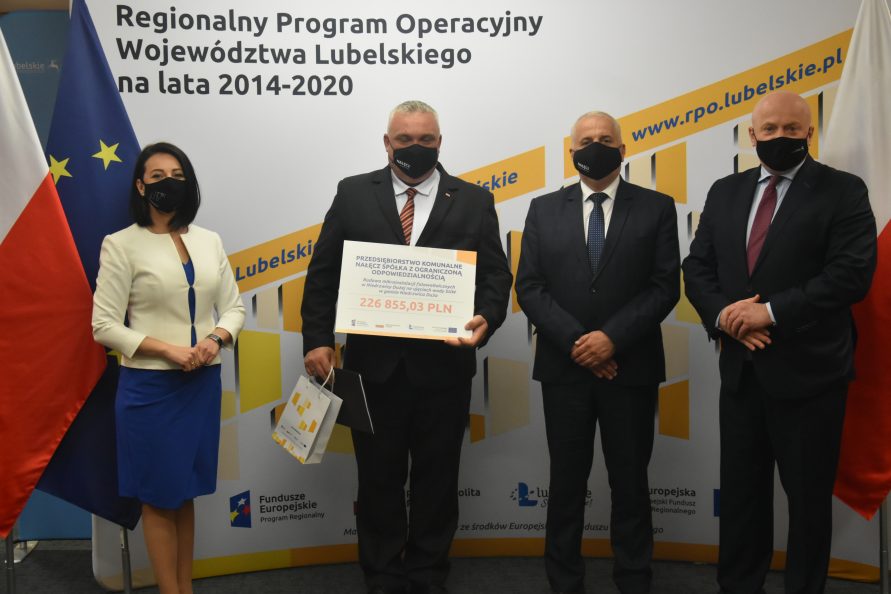 Na tle biało żółtej ścianki Regionalnego Programu Operacyjnego stoją od lewej: dyrektor LAWP Magdalena Filipek-Sobczak, po środku dwóch panów, przedstawiciele przedsiębiorstwa komunalnego Nałęcz sp. z o.o., pan po lewej trzyma w rękach symboliczny czek z wypisaną kwotą dofinansowania, z prawej marszałek Jarosław Stawiarski