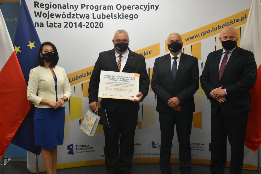 Na tle biało żółtej ścianki Regionalnego Programu Operacyjnego stoją od lewej: dyrektor LAWP Magdalena Filipek-Sobczak, po środku dwóch panów, przedstawiciele przedsiębiorstwa komunalnego Nałęcz sp. z o.o., pan po lewej trzyma w rękach symboliczny czek z wypisaną kwotą dofinansowania, z prawej marszałek Jarosław Stawiarski
