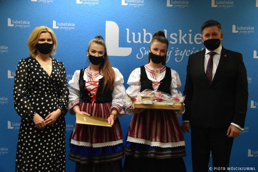 Na tle niebieskiej ścianki z napisami Lubelskie Smakuj życie stoją od lewej: dyrektor departamentu rolnictwa Ewa Szałachwiej, dwie laureatki w strojach ludowych trzymają nagrodę i pamiątkowy dyplom, członek zarządu Zdzisław Szwed