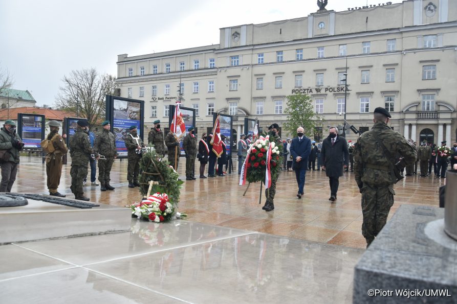 W dolnej części zdjęcia widok na grób nieznanego żołnierza. W centrum kadru żolnierz niosący białoczerwony wieniec, z tyłu idą członek zarządu Sebastian Trojak obok wicemarszałka Zbigniewa Wojciechowskiego. z lewej strony stoją poczty sztandarowe, w tle uczestnicy obchodów na tle budynku poczty polskiej przy placu Litewskim