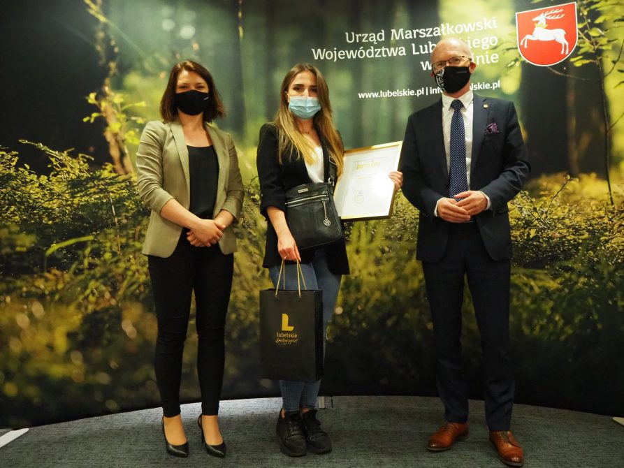 Po środku stoi laureatka konkursu fotograficznego, która trzyma dyplom i nagrodę. po prawej stoi członek zarządu Sebastian Trojak, po lewej wicedyrektor departamentu środowiska Emilia Flis