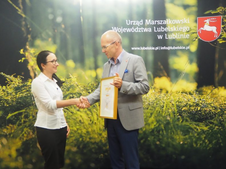 Członek zarządu Sebastian Trojak (z prawej) wręcza nagrodę laureatce konkursu instagramowego za miesiąc czerwiec. W tle ścianka przedstawiająca las. W górnym prawym rogu logotyp Urzędu Marszałkowskiego.