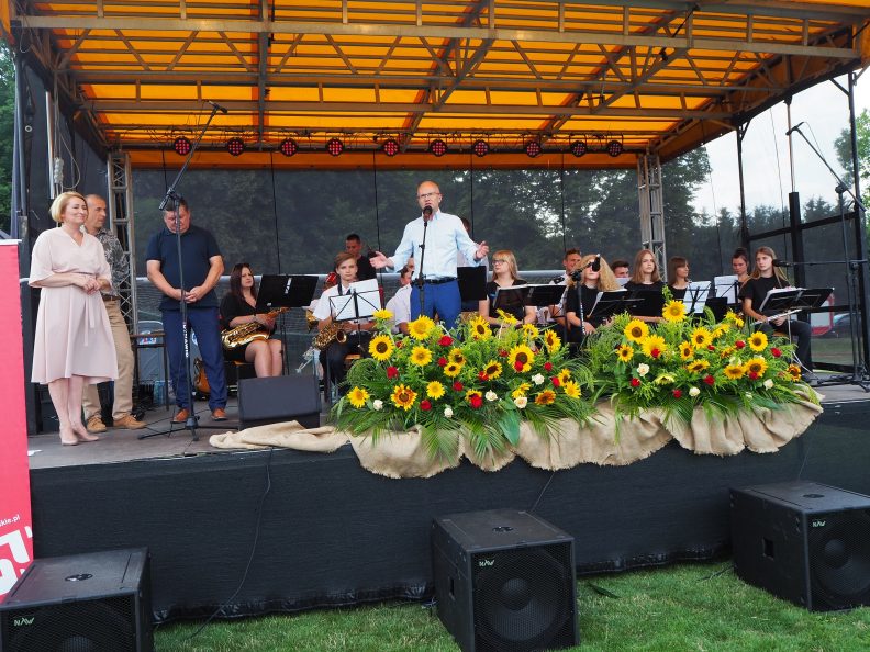 Na scenie po środku stoi Członek Zarządu Województwa Lubelskiego Sebastian Trojak, przed nim stoją bukiety słoneczników.