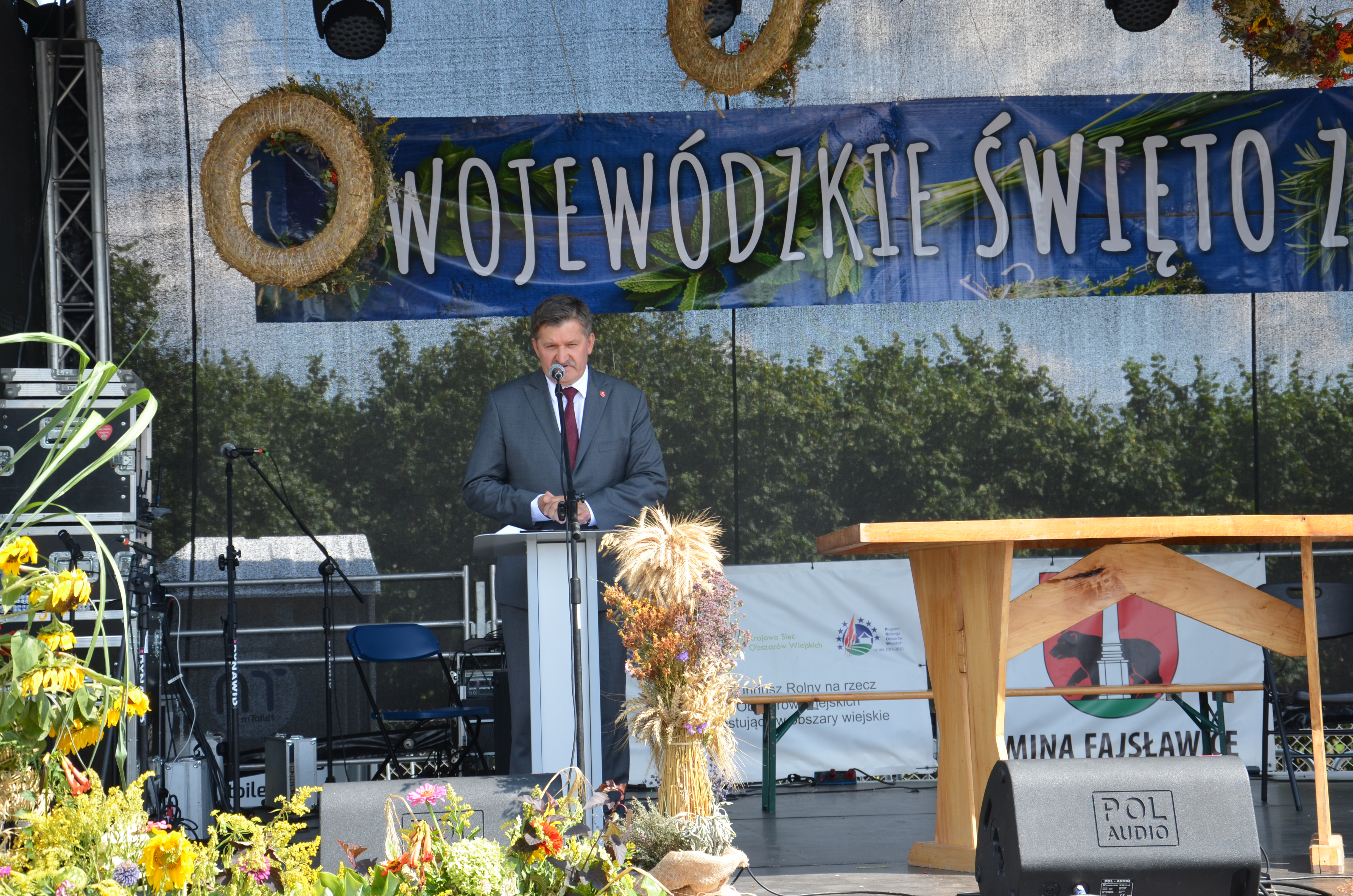 Na zdjęciu widoczny stojący na scenie, przy mównicy, Członek Zarządu Województwa Lubelskiego Zdzisław Szwed. W tle sceny widoczne korony drzew. Nad Panem Zdzisławem Szwed widoczny napis Wojewódzkie Święto Ziół. Przed nim stoi snopek zbóż ozdobiony ziołami i kwiatami