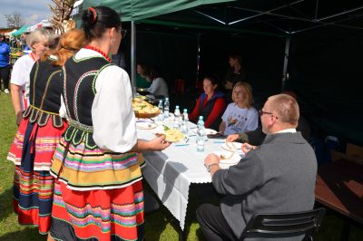 Komisja konkursowa siedząca przy stole nakrytym białym obrusem, Panie z Koła Gospodyń wiejskich ubrane w stroje ludowe prezentują dania konkursowe