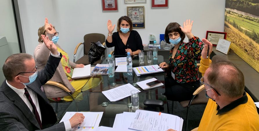 Przy szklanym stole siedzi pięcioro członków Kapituły konkursu, trzy panie i dwóch panów. Trzymają podniesione dłonie. Na stole leżą dokumenty.