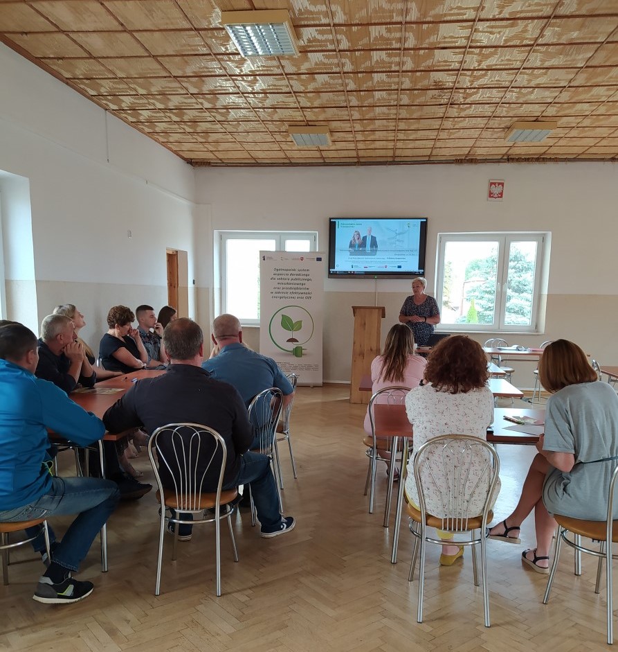 Zdjęcie przedstawiające stojącego Doradcę Energetycznego w sali w budynku, który przedstawia założenia programu Czyste Powietrze, któremu przysłuchują się siedzące na krzesłach przy stolikach osoby. Obok po lewej stronie doradcy znajduje się Baner Projektu Doradztwa Energetycznego.