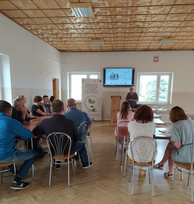 Zdjęcie przedstawiające stojącego Doradcę Energetycznego w sali w budynku, który przedstawia założenia programu Czyste Powietrze, któremu przysłuchują się siedzące na krzesłach przy stolikach osoby. Obok po lewej stronie doradcy znajduje się Baner Projektu Doradztwa Energetycznego.