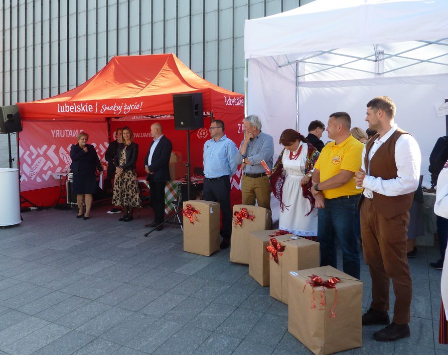 na zdjęciu widzimy conajmniej 8 osób, które ustawione są w rzędzie, Przed pięcioma osobami z prawej strony kadru stoją pudełka ozdobione kokardą. w tle namioty (biały i czerwony). Na czerwonym napis Lubelskie Smakuj życie.