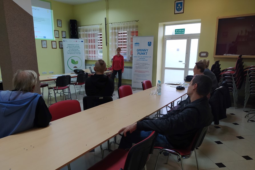 Zdjęcie przedstawiające stojącego Doradcę Energetycznego w sali w budynku, który przedstawia założenia programu Czyste Powietrze, któremu przysłuchują się siedzące na krzesłach przy stolikach osoby. Z tyłu doradcy znajduje się Baner Projektu Doradztwa Energetycznego a także baner zawierający m.in. napis Gminny Punkt.