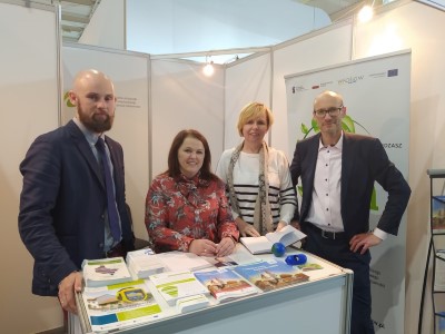 Zdjęcie przedstawiające 4 osoby stojące przy stoisku w pomieszczeniu. Na stoisku znajdują się foldery reklamowe. W tle znajdują się także banery reklamowe.