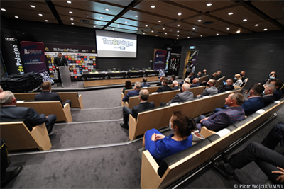 Widok na salę konferencyjną. na siedzeniach (tyłem do aparatu) siedzą uczestnicy konferencji. Z lewej przy mównicy na tle ścianki z logotypami sponorów Touru stoi prowadzący. Po środku stół prezydialny, nad nim ekran wyświetlający logo 78. Tour De Pologne