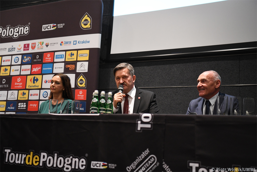 Przy stole prezydialnym, nakrytym czarną tkaniną z napisem Tour De Pologne siedzą trzy osoby, jedna kobieta (z lewej) i dwóch mężczyzn. Po środku członek zarządu Zdzisław Szwed trzymajacy mikrofon, z prawej Czesław Lang. W tle po lewej ścianka z logotypami sponsorów i partnerów wyścigu.