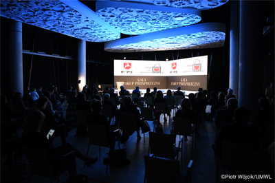 Widok na salę na której siedzą uczestnicy wydarzenia Gala Zakup Prospołeczny. Pomieszczenie wyciemnione, rozświetlony jest ekran wyświetlający logo ROPS i Zakup pro społeczny. Sufit podświetlony na niebiesko.