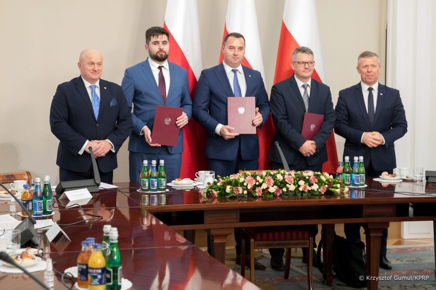 Na zdjęciu widać pięciu mężczyzn stojących za stołem na tle trzech flag Polski. trzech mężczyzn w środku trzyma teczki z aktem powołania.