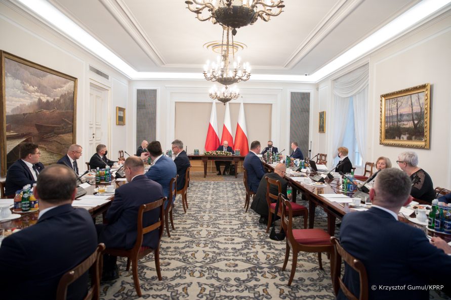 Widok na salę obrad. Stołu ustawione w podkowę. Przy nich siedzą członkowie Rady. W tle widać część prezydialną stołu, za nią trzy flagi polskie