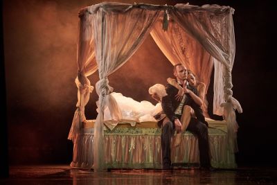 Plakat teatralny ukazujący mężczyznę i kobietę siedzących na łożu z baldachimem