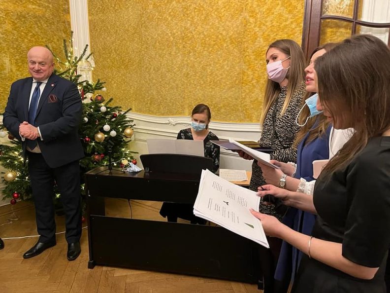 Zdjecie na którym po lewej stronie stoi marszałek województwa lubelskiego Jarosław Stawiarski a po prawej stronie stoją trzy kobiety, trzymające zapis nutowy i śpiewające kolędy, w tle choinka i muzyk grający na pianinie
