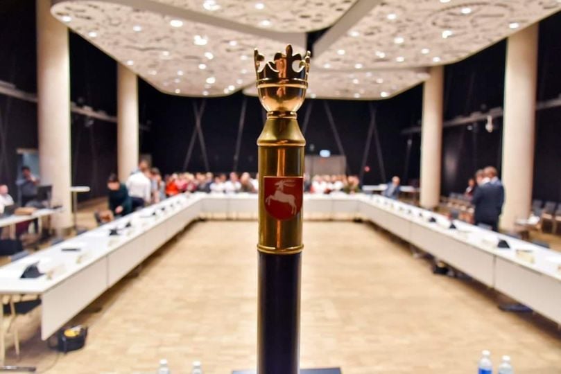 Obraz przedstawia salę posiedzeń Sejmiku Województwa Lubelskiego (w tle). Na pierwszym planie wybija się laska marszałkowska, a za nią widać w głębi stoły radnych województwa z kilkoma osobami, a na górze panele z oświetleniem. Zdjęcie jest w jasnej tonacji. Obraz w tle jest rozmyty.