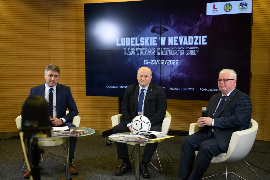 Zdjęcie przedstawiające trzech mężczyzn siedzących przy stoliku na którym leży położona piłka do gry w piłkę nożną