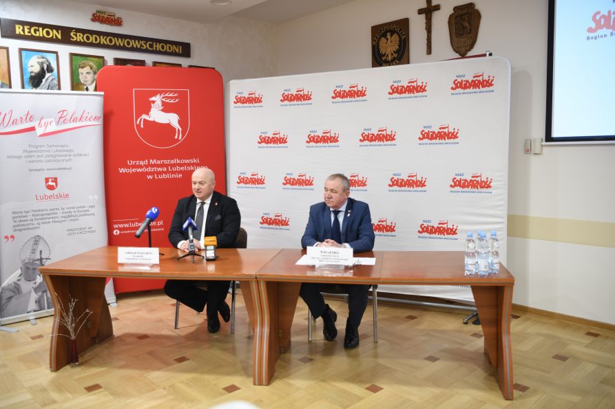 Zdjęcie przedstawiające Marszałka Województwa Lubelskiego Jarosława Stawiarskiego oraz Przewodniczącego NSZZ Solidarność Mariana Króla. Obaj panowie siedzą przy stole konferencyjnych, na którym ustawione są mikrofony. Za nimi znajdują się ścianki z logotypami województwa lubelskiego