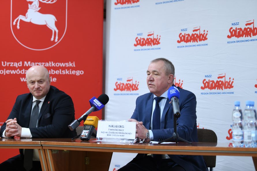 Zdjęcie przedstawiające Marszałka Województwa Lubelskiego Jarosława Stawiarskiego oraz Przewodniczącego NSZZ Solidarność Mariana Króla. Obaj panowie siedzą przy stole konferencyjnych, na którym ustawione są mikrofony. Za nimi znajdują się ścianki z logotypami województwa lubelskiego