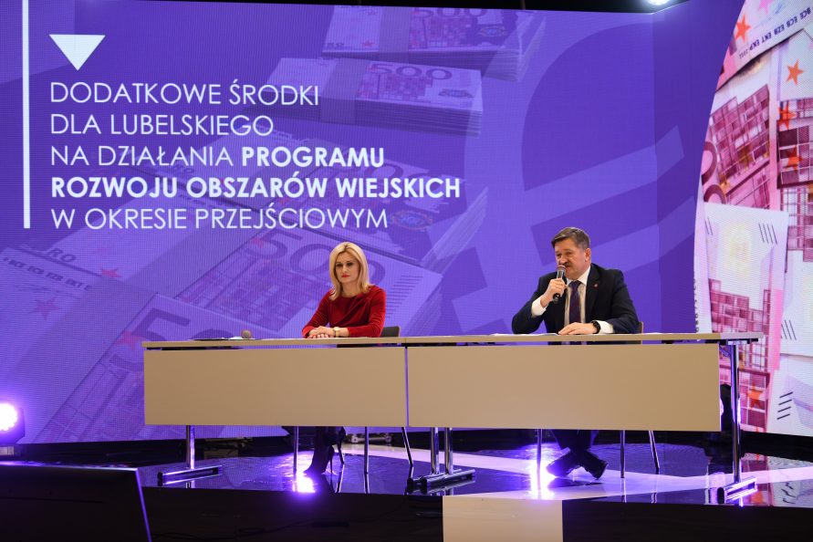 Zdjęcie przedstawiające dwie osoby, kobietę i mężczyznę siedzących przy stole konferencyjnym i udzielających wywiadu. Za nimi w tle znajduje się ogromny telebim z napisem Dodatkowe środki dla lubelskiego na działania Programu Rozwoju Obszarów wiejskich w okresie przejściowym