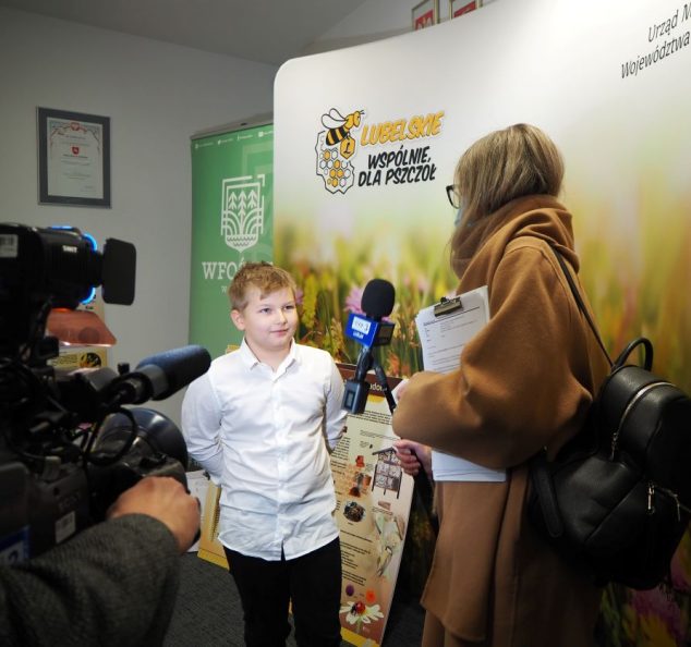 do Na zdjęciu laureat pierwszego miejsca Jan Rodziewicz udziela wywiadu reporterce TVP 3 Lublin na tle ścianki promocyjnej kampanii "Lubelskie - wspólnie dla pszczół". Obok, z lewej, fragment kamery telewizyjnej