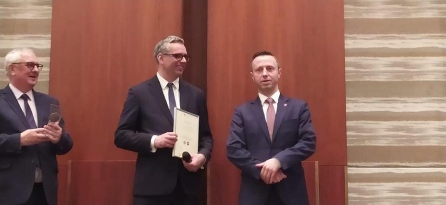 Zdjęcie przedstawiające trzech mężczyzn stojących przodem do kamery, z czego mężczyzna stojący w środku trzyma w rękach dyplom