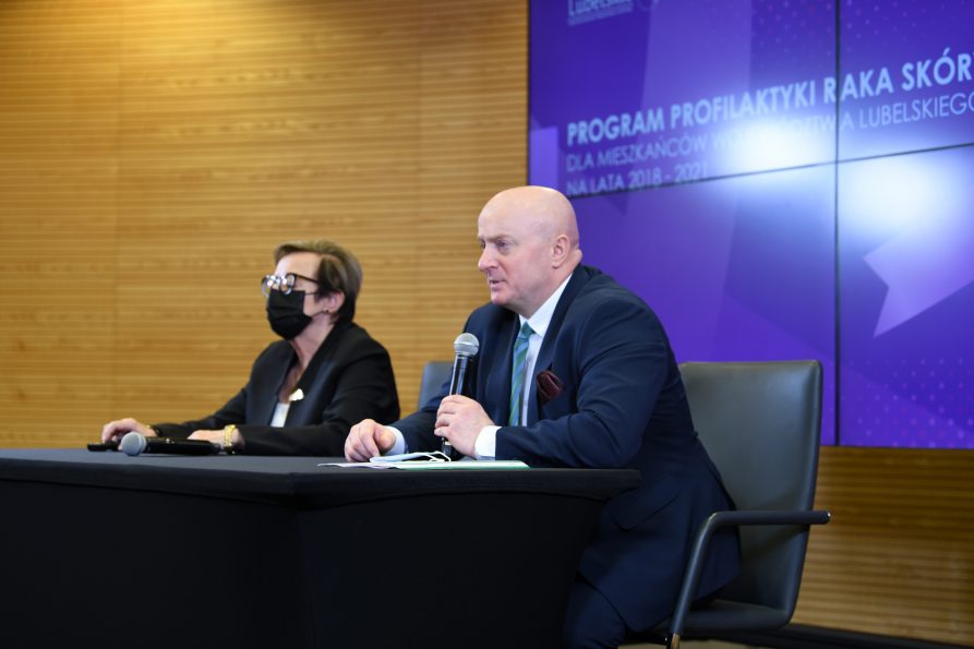 Zdjęcie przedstawiające dwie osoby, kobietę i mężczyznę, którzy siedzą przy stole konferencyjnym. Kobieta ma założoną maseczkę a mężczyzna trzyma w ręce mikrofon i udziela wypowiedzi. Z tyłu w tle znajduje się duży telebim z logotypem programu profilaktyki raka skóry.