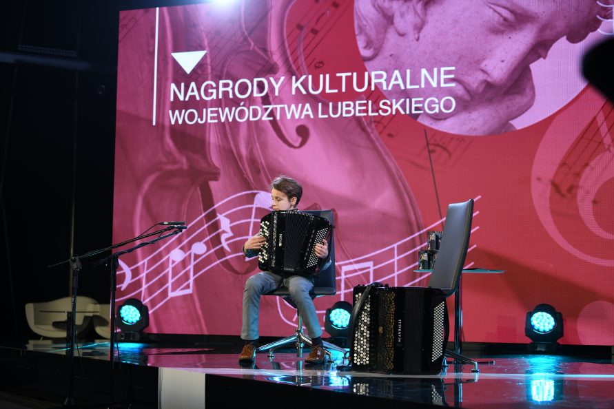 Zdjęcie przedstawiające chłopca siedzącego na scenie i grającego na akordeonie