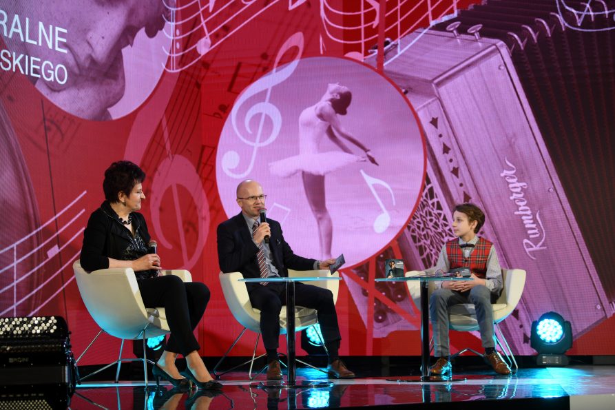 Zdjęcie przedstawiające trzy osoby siedzące na scenie kobietę, mężczyznę i chłopca