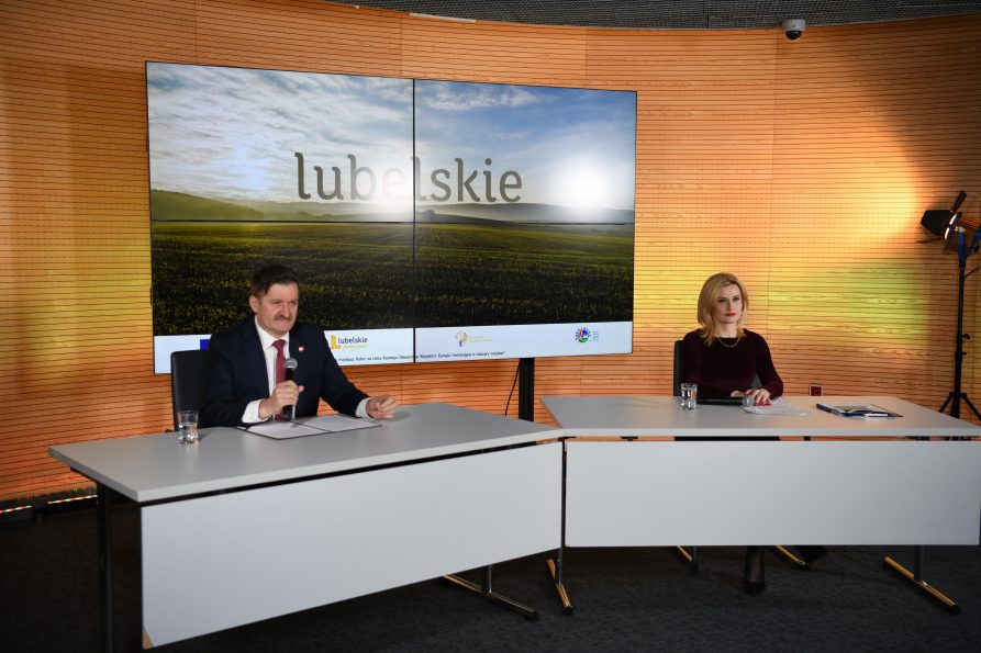 Zdjęcie przedstawia studio nagraniowe w którym siedzą za stołem kobieta i mężczyzna i udzielają wywiadu. Za nimi w tle znajaduje się duży ekran wyświetlający zdjęcie zielonego pola i napis na nim lubelskie