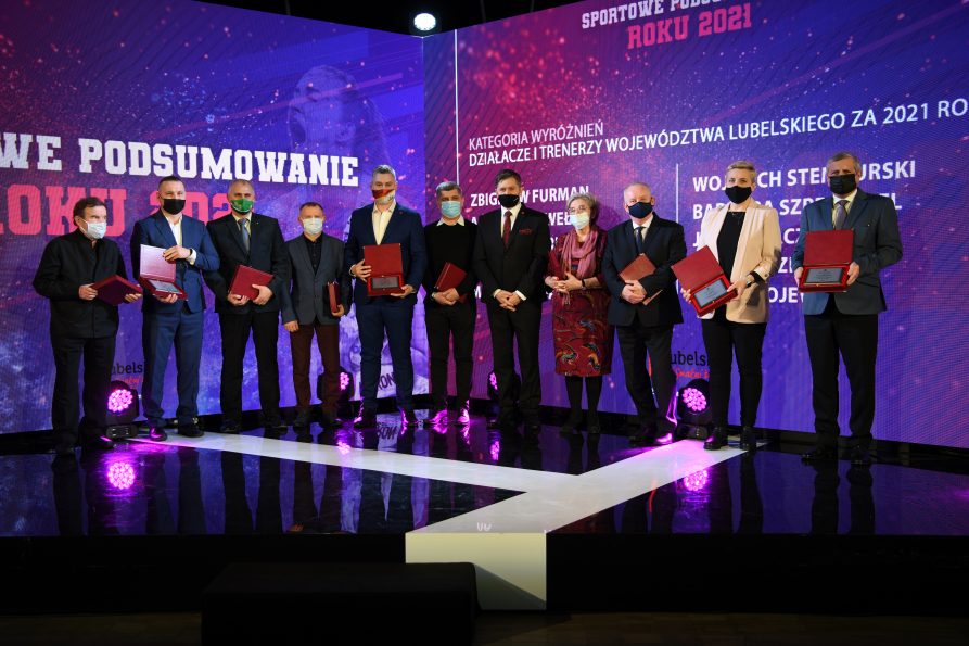 Zdjęcie przedstawia grupę osób kobiet i męzczyzn, którzy stoją na dużej scenie i trzymają dyplomy, z niami znajduje się ogromny ekran wyswietlajacy napis Sportowe podsumowanie roku 2021