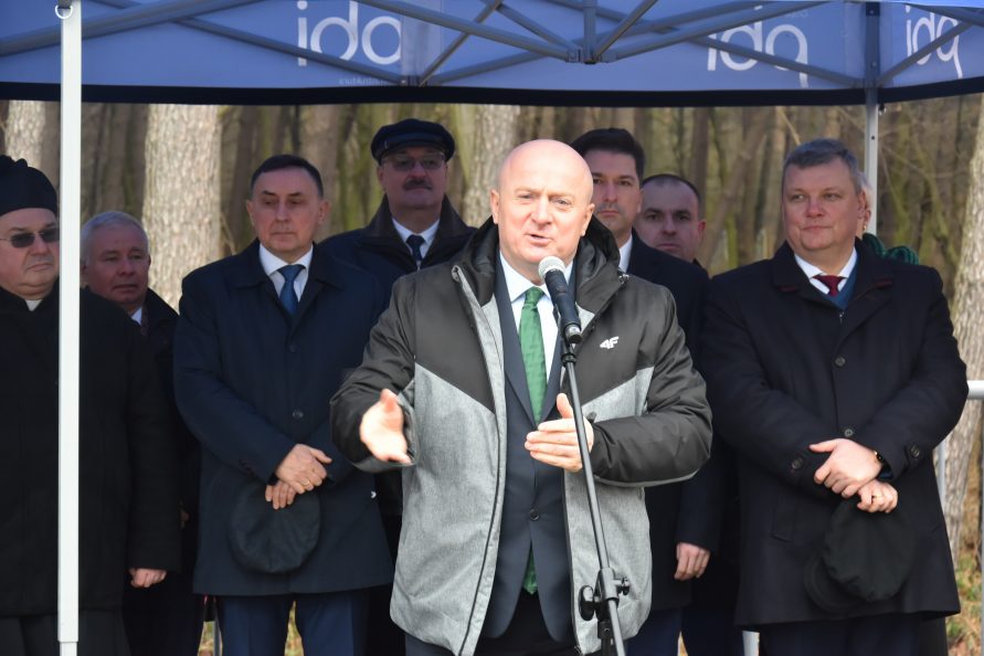 Zdjęcie przedstawia mężczynę stojącego przy mikrofonie pod namiotem na dworze, udzielającego wypowiedzi. W tle stoi grupa mężczyzn i przysłuchuje się wypowiedzi.