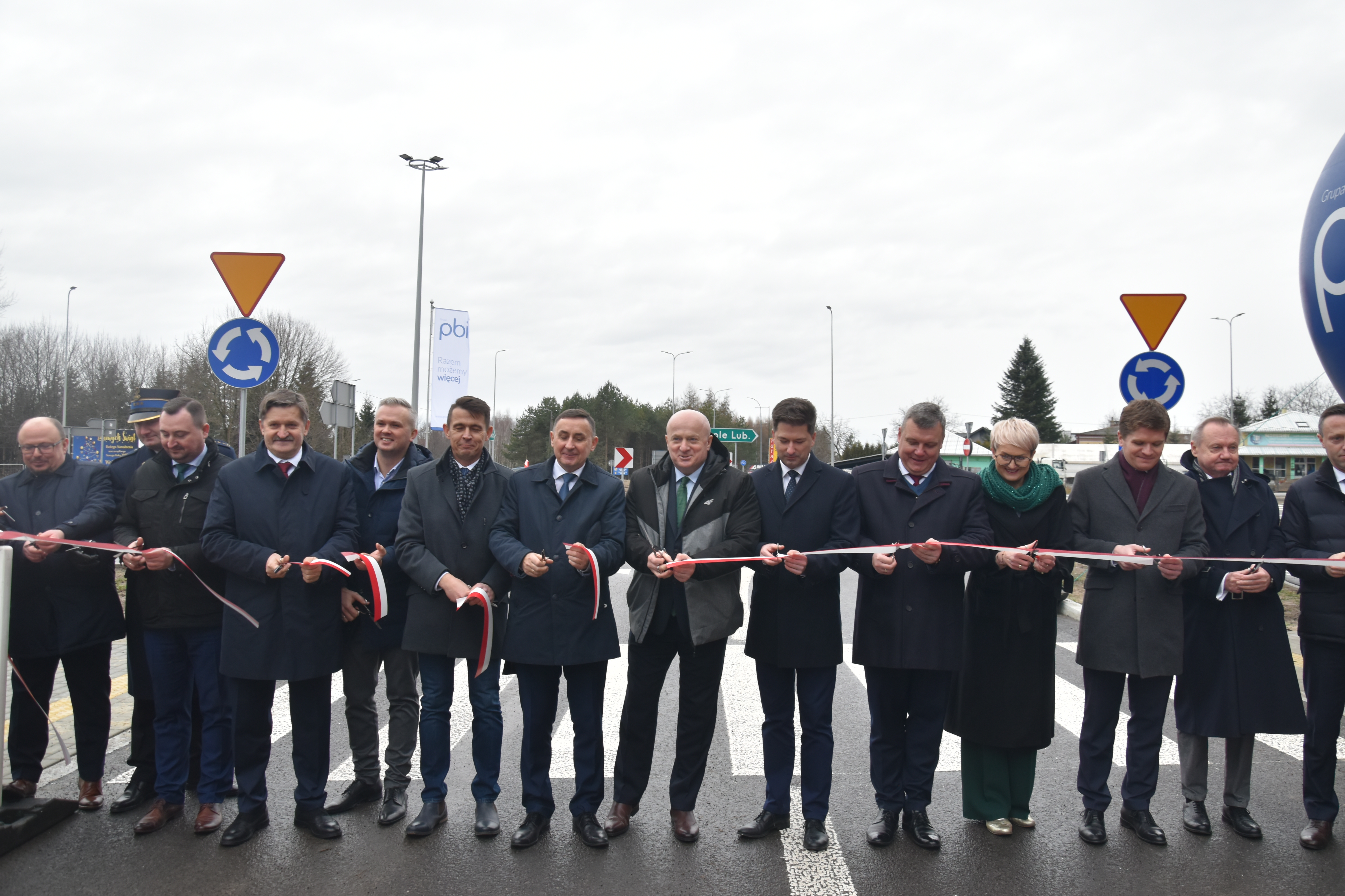 Grupa mężczyzn stoi obok siebie, wszyscy trzymaja w rekach jedną długa wstęgę, która przetną jako symbol otwarcia nowej drogi