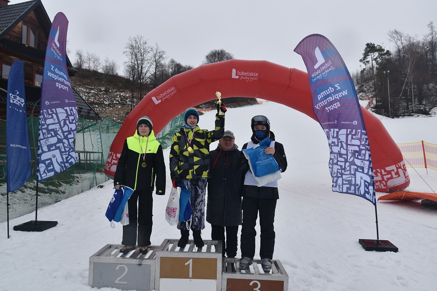Zdjęcie przedstawia podium dla laureatów w wyróżnionym stopniem na miejsce pierwsze, drugie i trzecie. Na każdym ze stopni stoi chłopcie który zajął adekwatne miejsce, wszyscy trzej mają medale a jeden z nich trzyma puchar. Za nimi stoi mężczyzna, który wręczył im nagrody, wszystko odbywa się na zasnieżonym stoku.