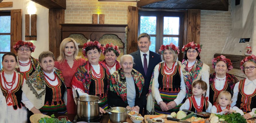 Zdjęcie grupowe pań z koła gospodyń wiejskich ubranych w stroje ludowe i wianki na głowach. Wśród pań stoi Członek Zarządu Pan Zdzisław Szwed oraz Dyrektor Departamentu Rolnictwa i Rozwoju Obszarów Wiejskich. W tle wiejska izba.