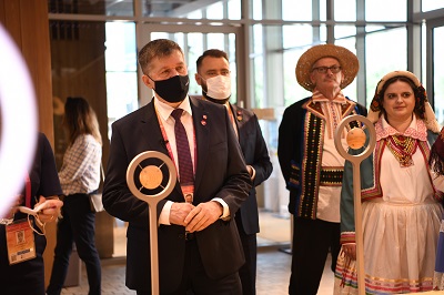 Zdjecie przedstawia mężczyznę stojącego przed mikrofonem w maseczce a obok niego tancerze w strojach ludowych