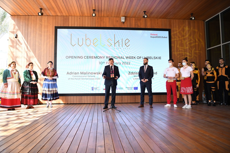 Zdjecie przedstawiające dwóch męzczyzn którzy stoją na dużej sali przy mirofonach i udzielają wypowiedzi, obok nich stoją czlonkowie zespołu tańca w ludowych strojach