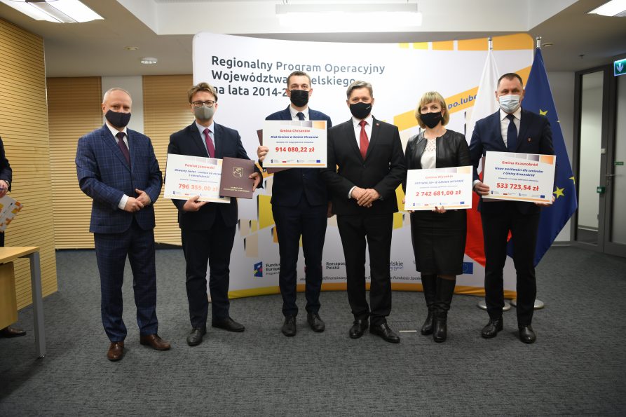 Na zdjęciu znajduje się kilka osób mężczyzni i jedna kobieta i prezentują trzymając w rękach symboliczne umowy na dofinasowanie