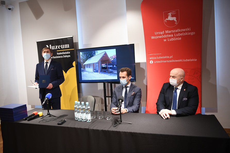 Zdjęcie przedstawiające trzech mężczyzn, dwóch z nich siedzi przy stoliku z ustawionymi przed nimi mikrofonami, trzeci stoi w obrębie stołu i udziela wypowiedzi